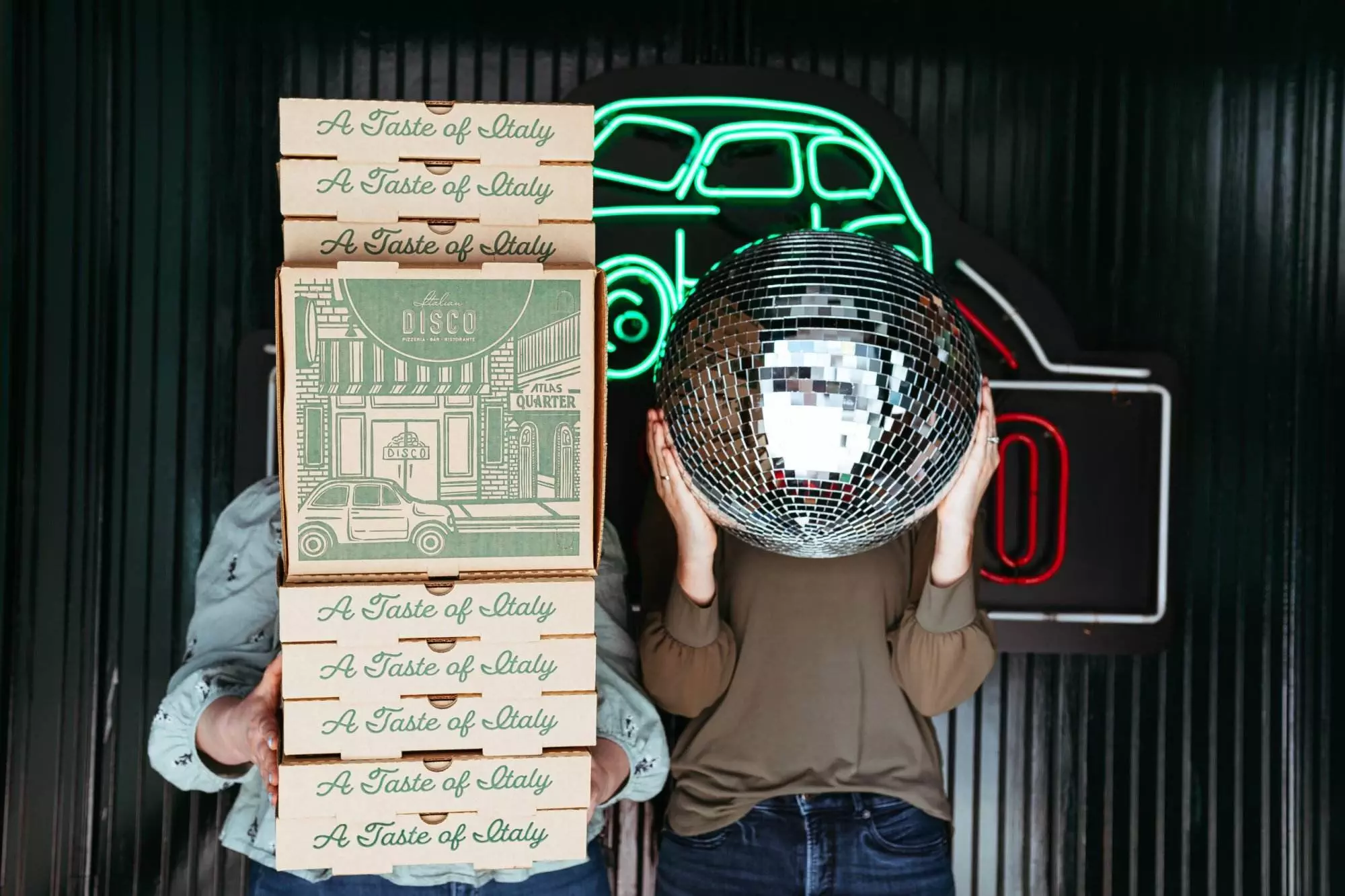 Image of two people, one is holding stack of pizza boxes and the other is holding a pizza box